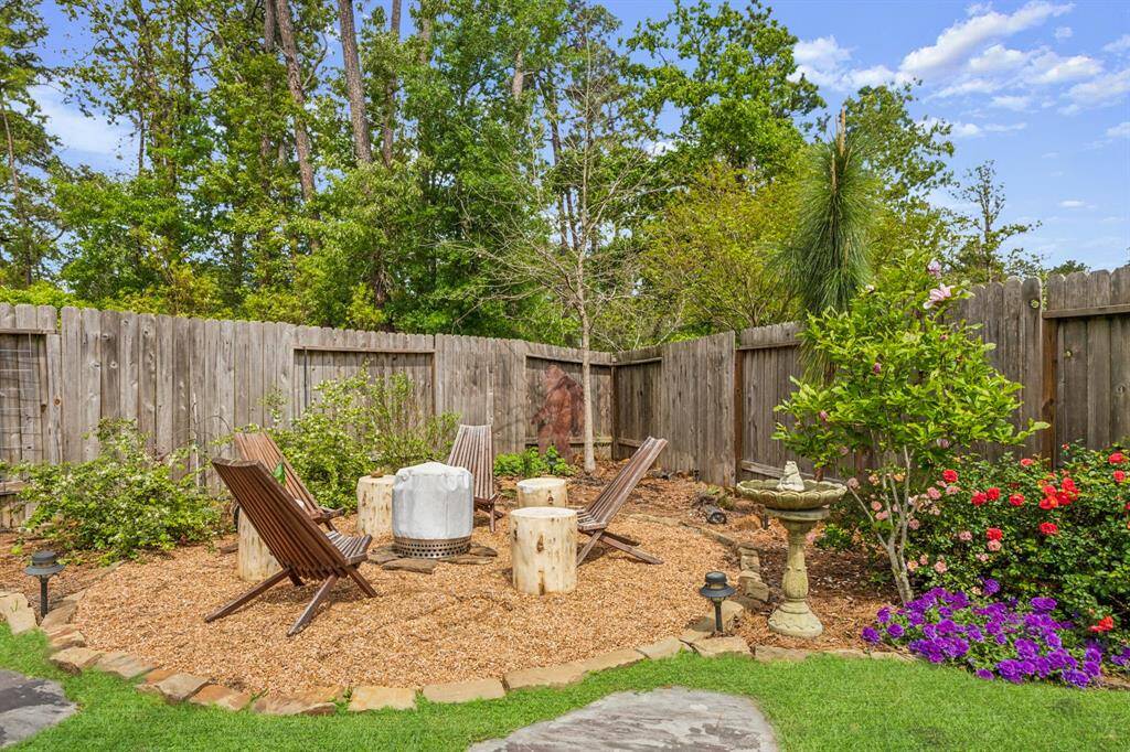 This backyard features a cozy seating area with wooden chairs around a fire pit, set on a mulched surface. It's surrounded by a wooden fence and lush greenery, including flowering plants and shrubs, creating a private, tranquil retreat. A birdbath adds a charming touch.