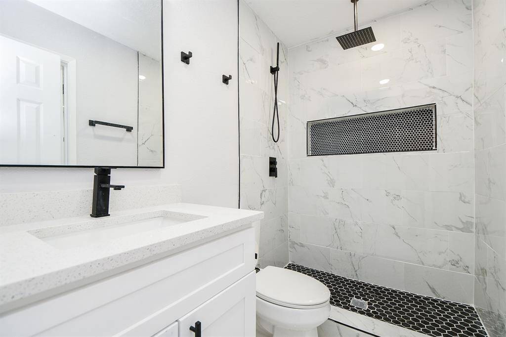The primary bedroom's modern bathroom features a sleek vanity with a black faucet, a large mirror, and a spacious marble-tiled shower with a rainfall showerhead. The dark hexagonal floor tiles and recessed wall shelf add a stylish touch.