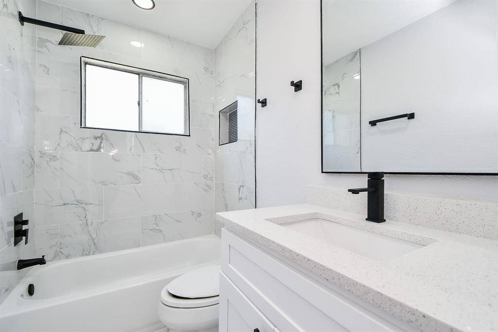 This hall bath upstairs features a modern design with white marble-style tiles, a sleek black faucet, and fixtures. It includes a bathtub with an overhead shower, a large mirror, and a window for natural light.