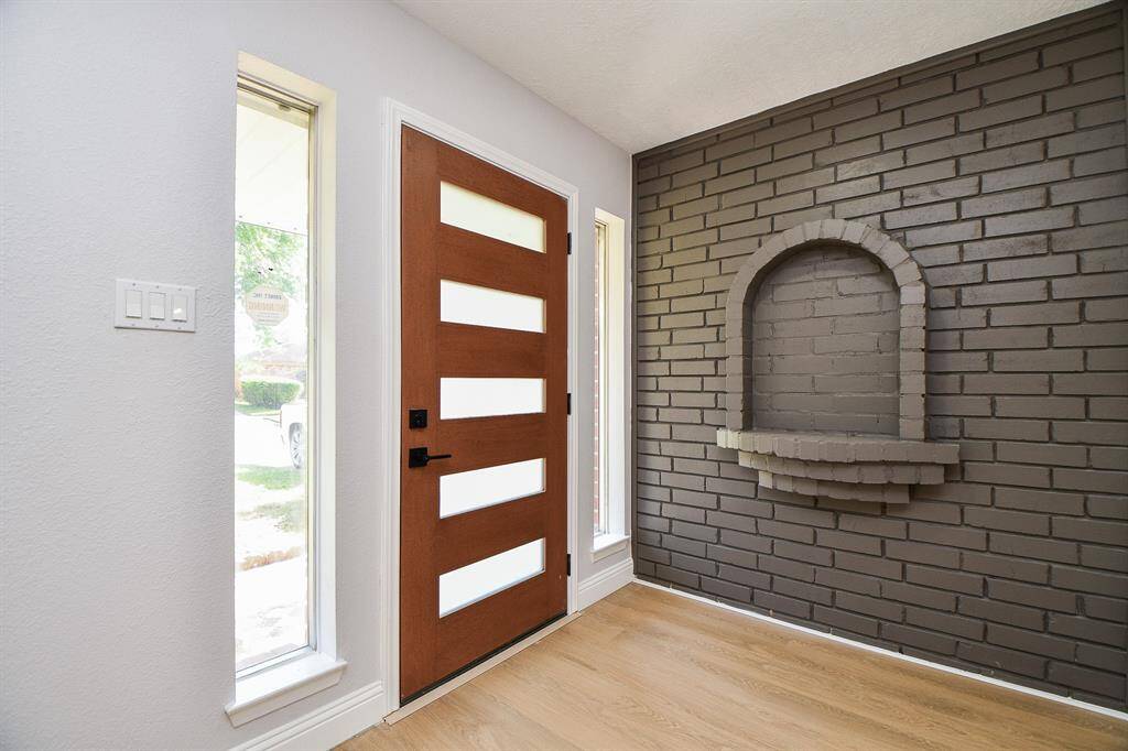 Walk into your home through this modern wooden door with horizontal glass panels, flanked by tall windows for ample natural light. The interior showcases a stylish gray brick accent wall with a built-in niche, complemented by light laminate wood flooring.
