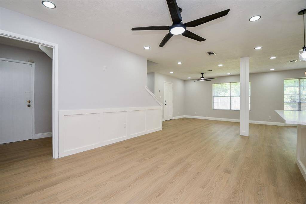 Imagine yourself in this spacious living area that features modern recessed lighting, ceiling fans, and large windows for natural light. The open floor plan includes light wood flooring and wall wood accents, offering a contemporary and inviting atmosphere.