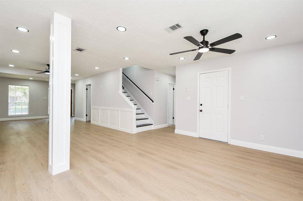 A modern, open-concept living space with light neutral walls. It features a ceiling fan, a staircase with a sleek railing, and abundant natural light from large windows.