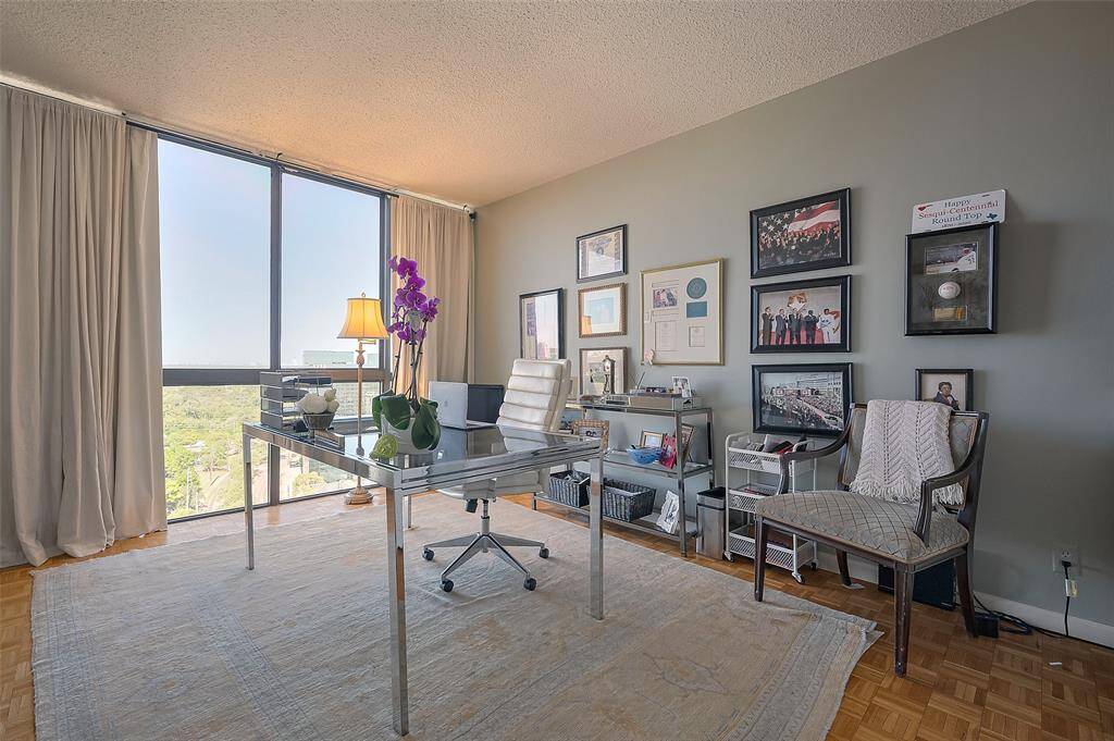 Bright home office with large windows offering scenic views, modern glass desk, and ample wall space featuring framed artwork. Cozy and functional workspace.