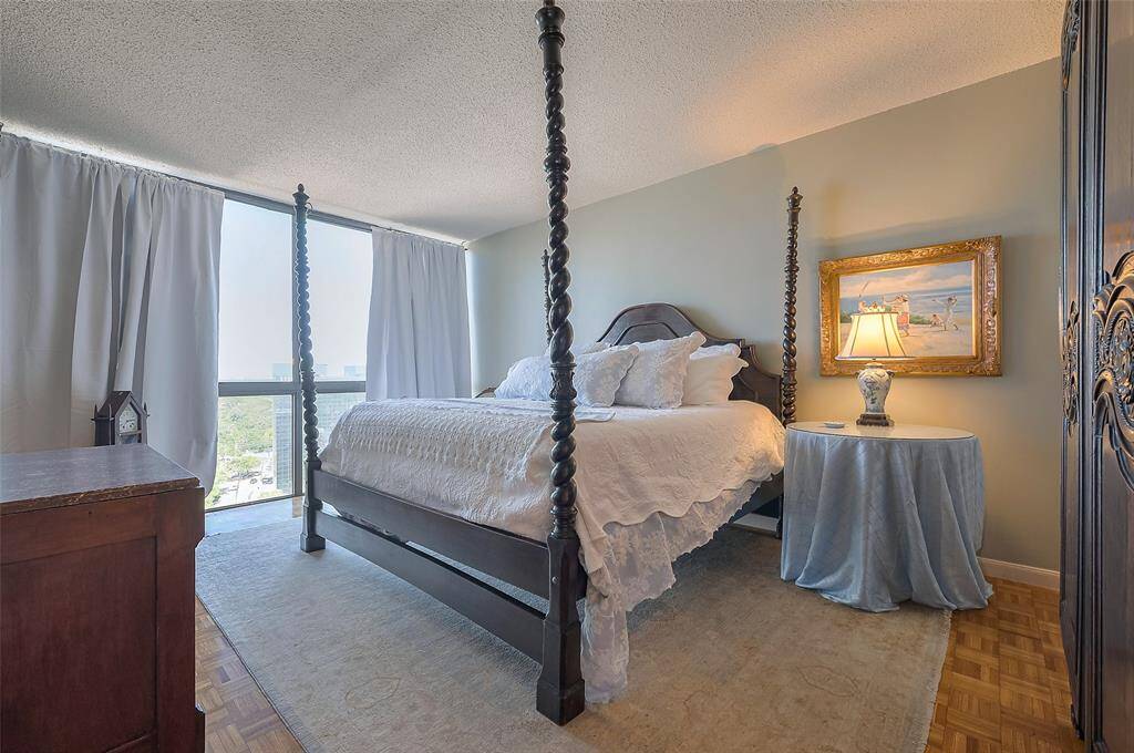 Bright bedroom featuring a stylish four-poster bed, elegant decor, and large windows with curtains, offering a city view. The room includes a cozy side table with a lamp and a decorative painting, creating a warm and inviting atmosphere.