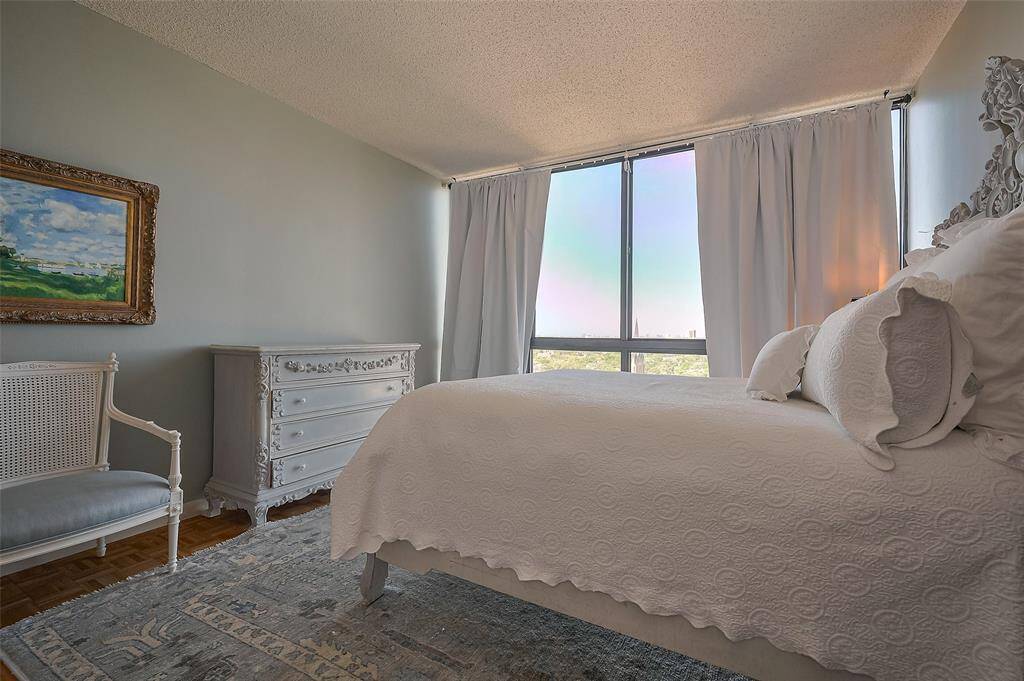 This bedroom features a serene, elegant design with soft gray walls, a large window with curtains, and an abundance of natural light. It includes a white ornate dresser, a matching chair, and a plush bed, creating a cozy and inviting atmosphere.