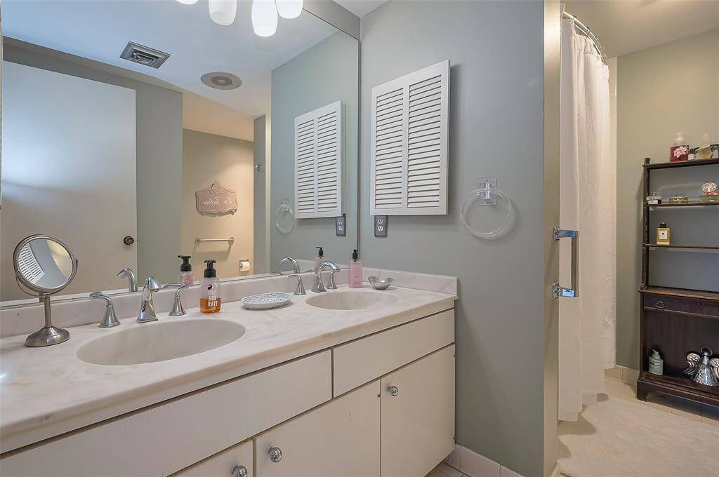 This bathroom features a double sink vanity with a large mirror, soft lighting, and ample cabinet storage. It includes a shower with a curtain, a wall-mounted towel ring, and a shelf for additional storage.