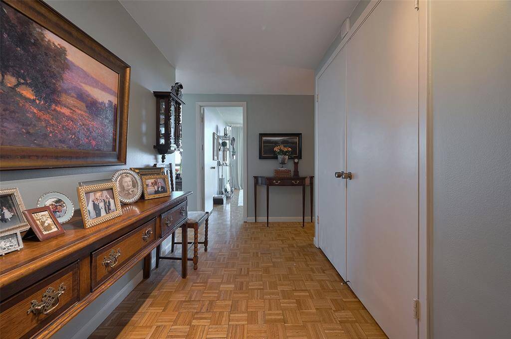 a bright hallway with parquet flooring and neutral walls. It features decorative elements like framed photos on a wooden console and a painting.