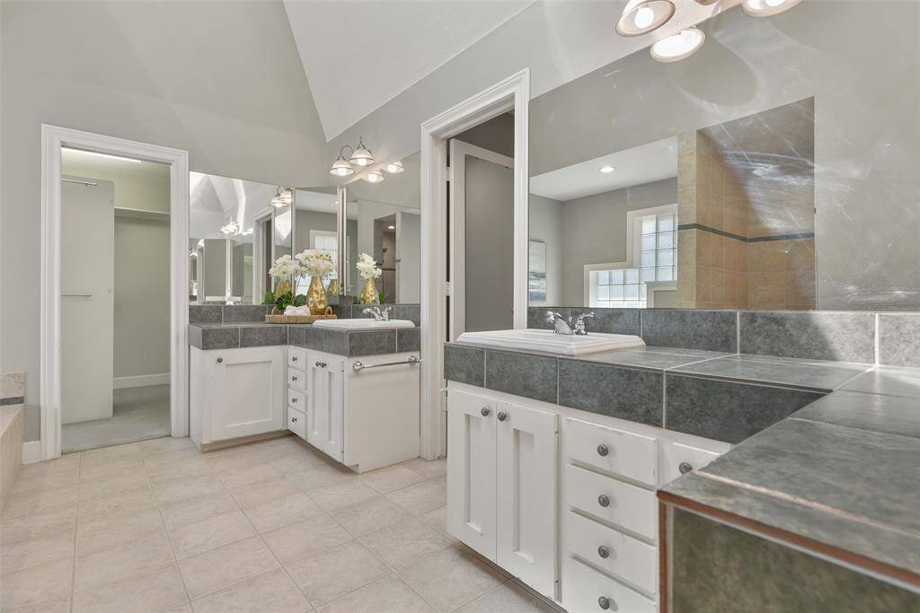 Relaxation awaits in this master bathroom, which features separate his-and-hers vanities with tile counters, dual rectangular basin sinks, white cabinetry, a large jetted soaker tub, a zero-entry walk-in shower with a handheld showerhead, and a roomy walk-in closet with a built-in dresser.
