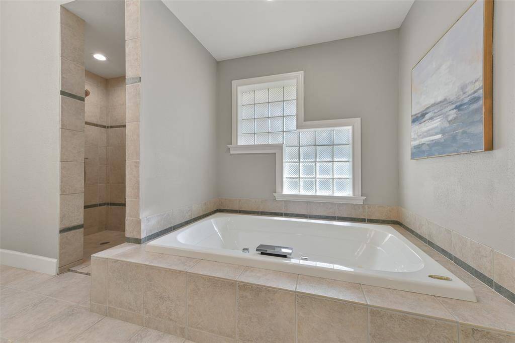 Relaxation awaits in this master bathroom, which features separate his-and-hers vanities with tile counters, dual rectangular basin sinks, white cabinetry, a large jetted soaker tub, a zero-entry walk-in shower with a handheld showerhead, and a roomy walk-in closet with a built-in dresser.