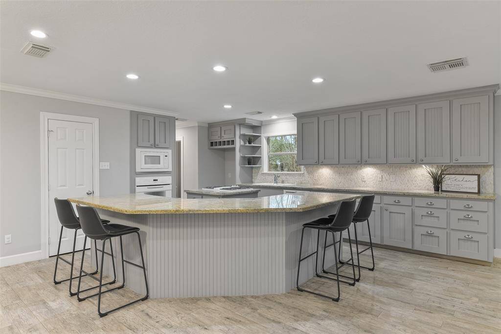 A semi-circular wrap-around breakfast bar with ample seating space defines this open-concept island kitchen with gleaming granite countertops, abundant grey cabinetry with silver hardware, and a beautifully patterned stone tile backsplash.
