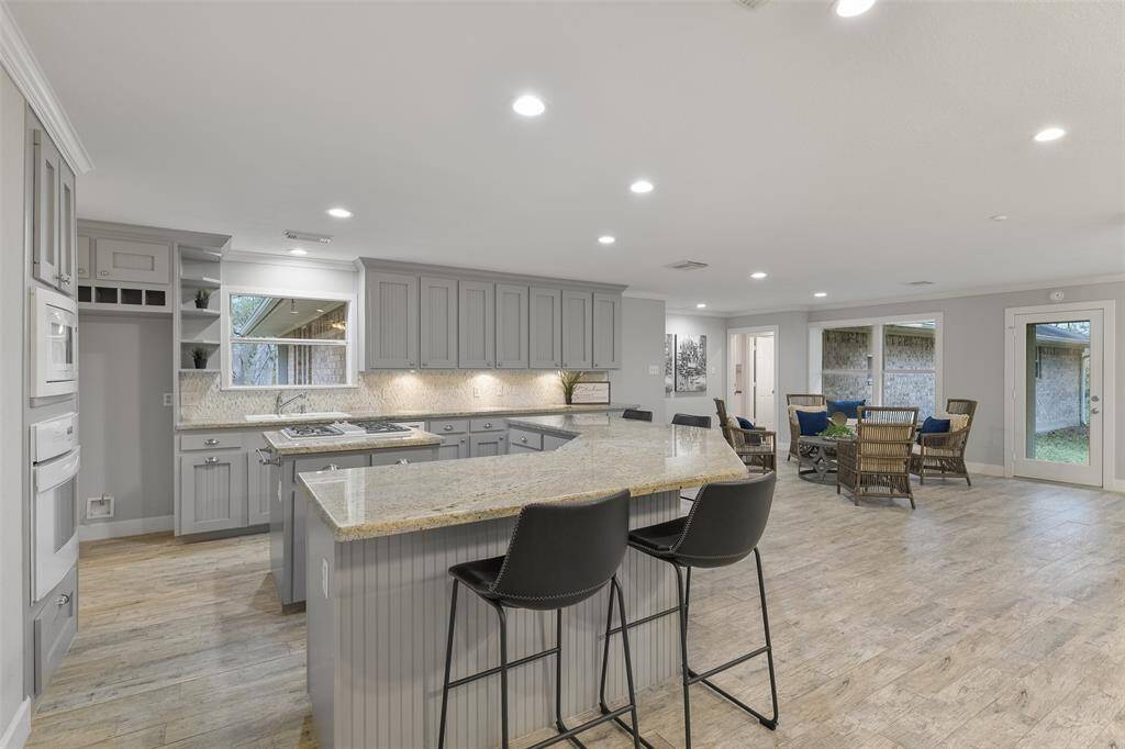 A semi-circular wrap-around breakfast bar with ample seating space defines this open-concept island kitchen with gleaming granite countertops, abundant grey cabinetry with silver hardware, and a beautifully patterned stone tile backsplash.
