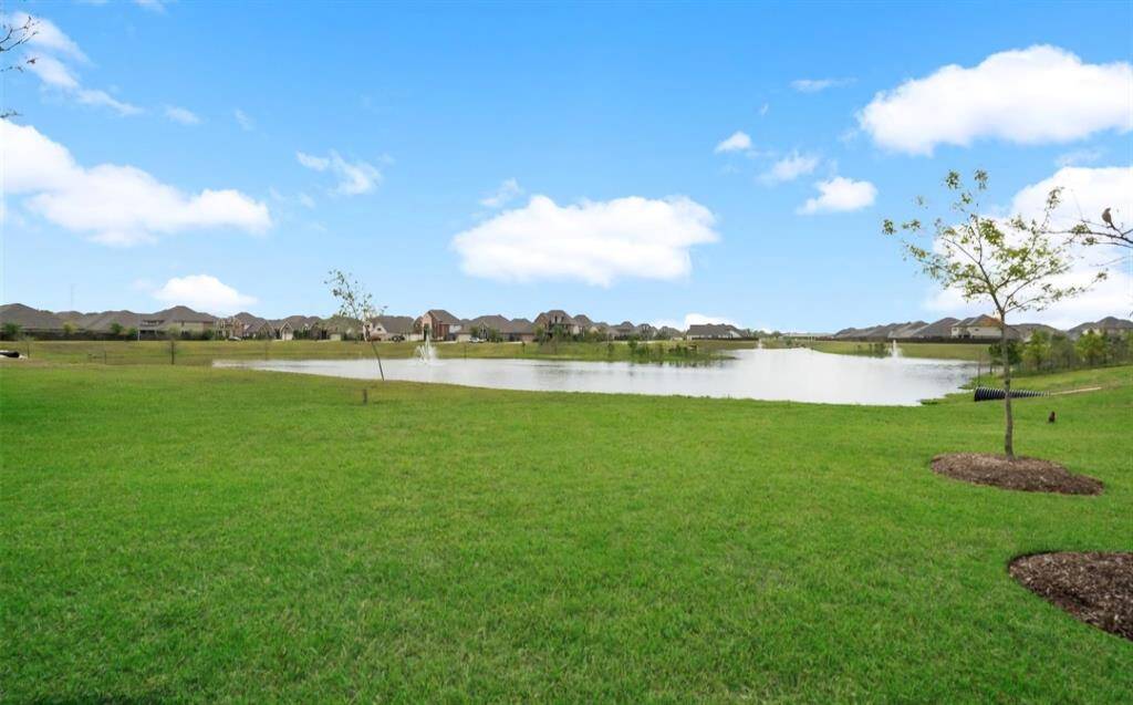Serene lakeside view with calm waters and a fountain. In the background, a row of houses is visible, surrounded by trees, offering a peaceful neighborhood setting.