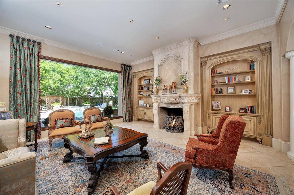 The elegant formal living room located off the foyer features travertine tile flooring, 11” baseboards, faux finished walls, carved crown molding, recessed and art lighting, surround sound, custom arched wood-stained built-ins with lighted upper shelving with lower cabinetry, and a carved limestone fireplace and overmantel. A double-butted wall of glass overlooks the stunning backyard and fills this space with natural light.