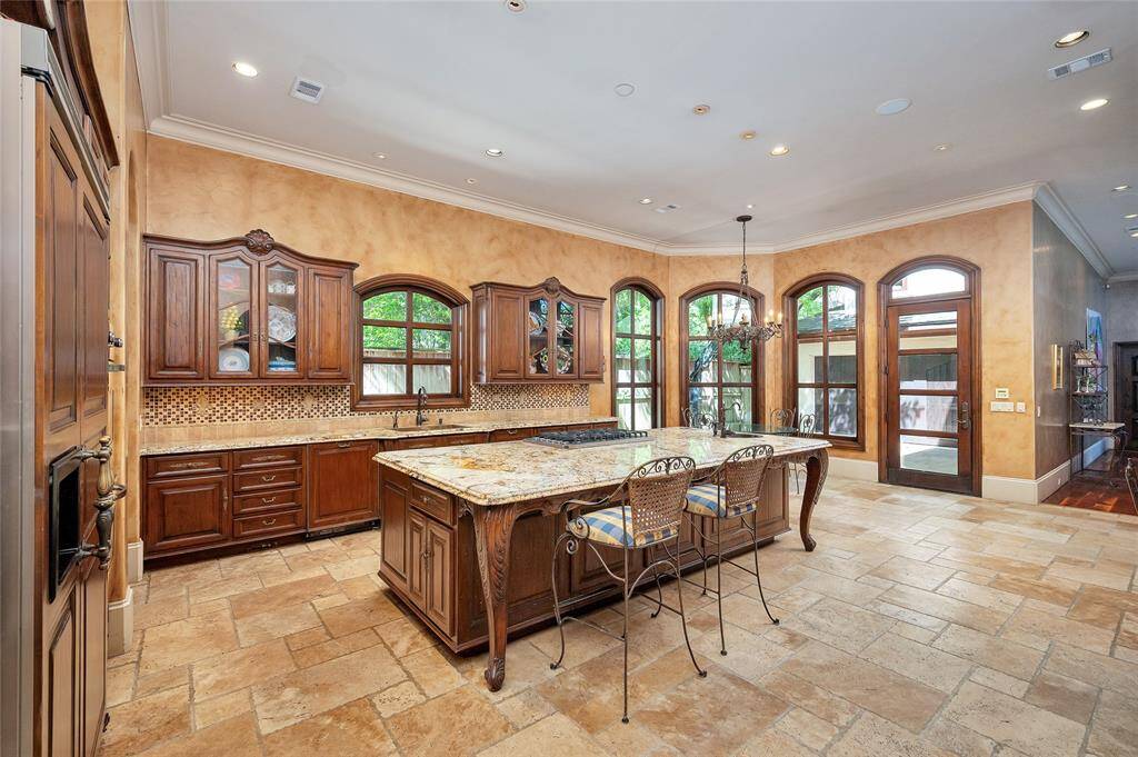 The chef’s kitchen boasts a large central island with seating, custom furniture-style wood-stained cabinetry, glass front upper cabinets, granite countertops, mosaic tile backsplash with fleur-de-lis and rosette medallions, stainless steel and paneled appliances, custom Italian hardware, and under-cabinet lighting.