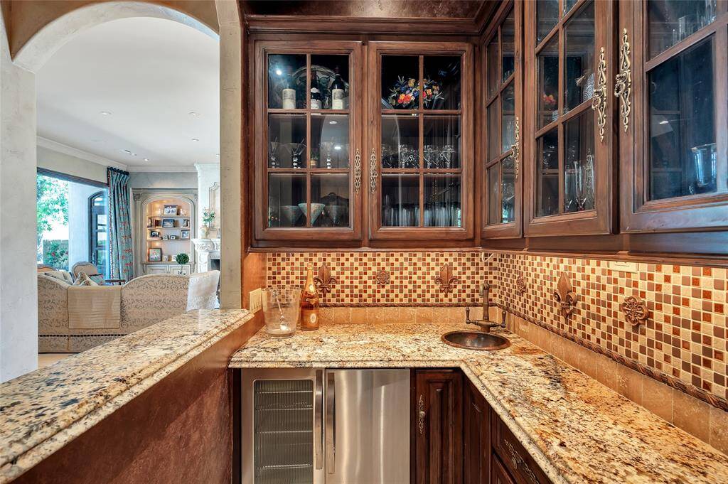 Accessible from the kitchen, the wet bar features a rich wood-stained built-in cabinetry, glass front upper cabinets, granite countertops, a mosaic tile backsplash with fleur-de-lis and rosette medallions, hammered copper bar sink, stainless Viking beverage fridge and ice maker, and a barrel vault ceiling. An arched serving window with art lighting and a raised granite bar makes entertaining and serving guests a breeze.