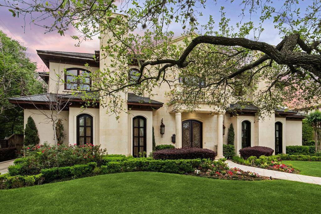 The grand facade features a stucco exterior, arched windows, twin chimneys, and a Juliet balcony. The manicured lawn and lush landscaping enhance its curb appeal, while a curved pathway leads to a covered entry with stately French doors.