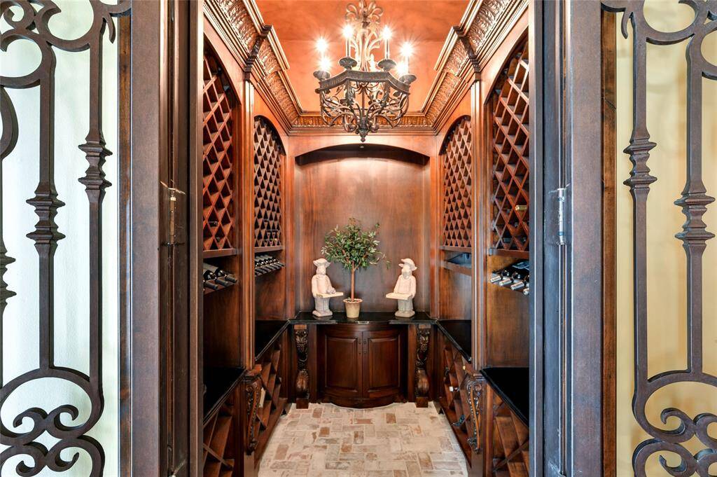 Ideal for wine enthusiasts, the sunken wine room with wrought iron French door entry includes brick herringbone flooring, custom wood-stained built-ins with intricate moldings and accent lighting, multiple wine racks, black granite countertops, and wrought-iron chandelier lighting.