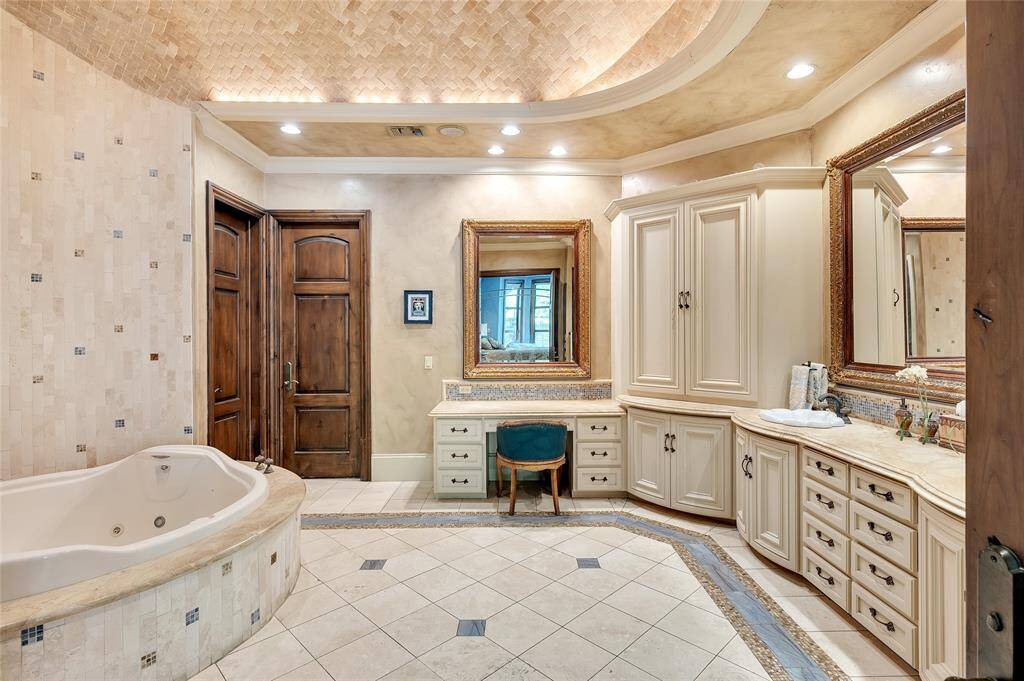 The spa-like primary bathroom boasts a round jetted tub with Vita Bath controls, an accent wall with travertine tiles inlayed with marble and glass accents, and a custom wooden built-in double-vanity with a separate sitting area, marble countertops, and gold-framed vanity mirrors. Additional features include travertine tile flooring with blue marble and bronze mosaic tile inlays, a matching backsplash, and barrel-vault ceiling clad in herringbone travertine tile with accent lighting.