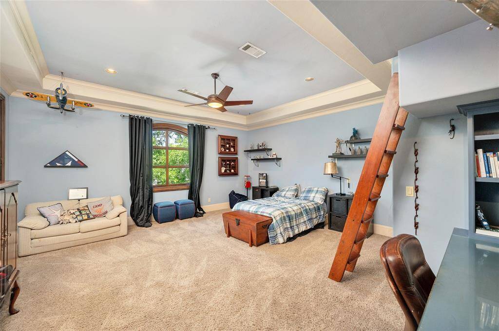 This large secondary bedroom with an en-suite bathroom and walk-in closet features plush neutral carpet, 11” baseboards, soft blue walls, tray ceiling, arched window with draperies, recessed lighting, and a decorative ceiling fan. Bonus features include access to the exterior balcony, a built-in desk, recessed built-in bookshelves, and a ladder to a in-wall play tunnel system.
