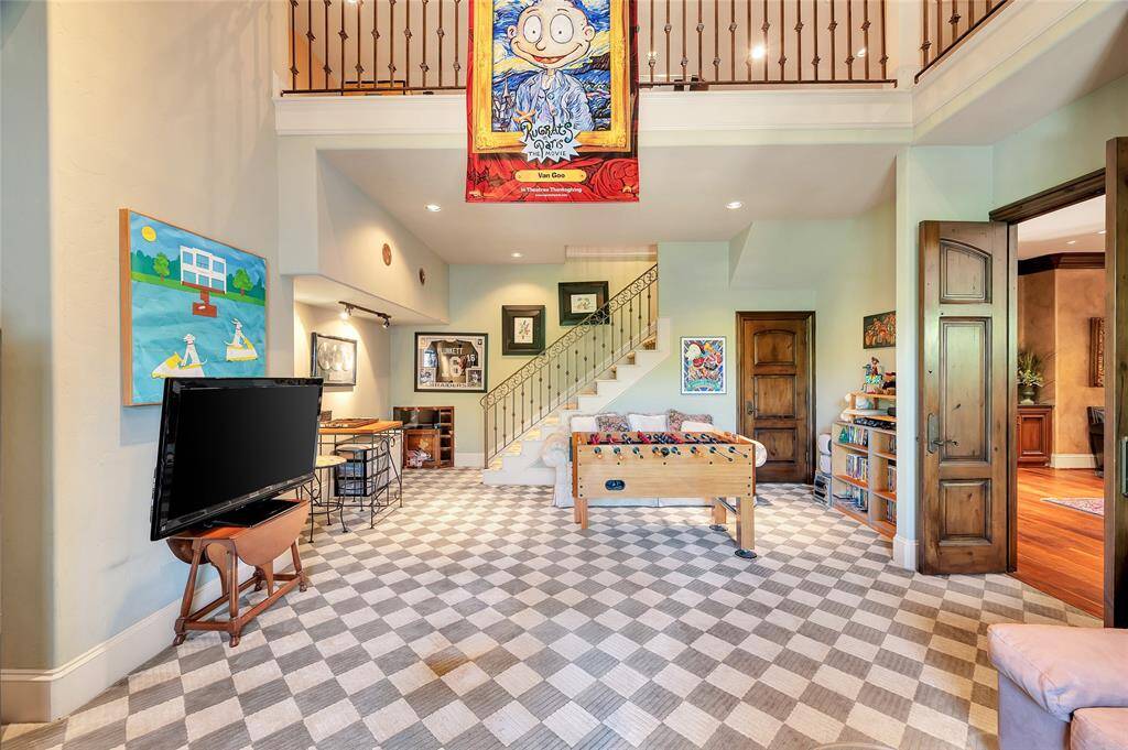 This vibrant 2-story children’s gameroom boasts checkered carpet flooring, double-story ceilings, a large storage closet, glass-paneled French doors that open to the exterior balcony, a staircase with wrought iron railing that leads to the upper level, and a play tunnel system with nautical brass window lookouts.