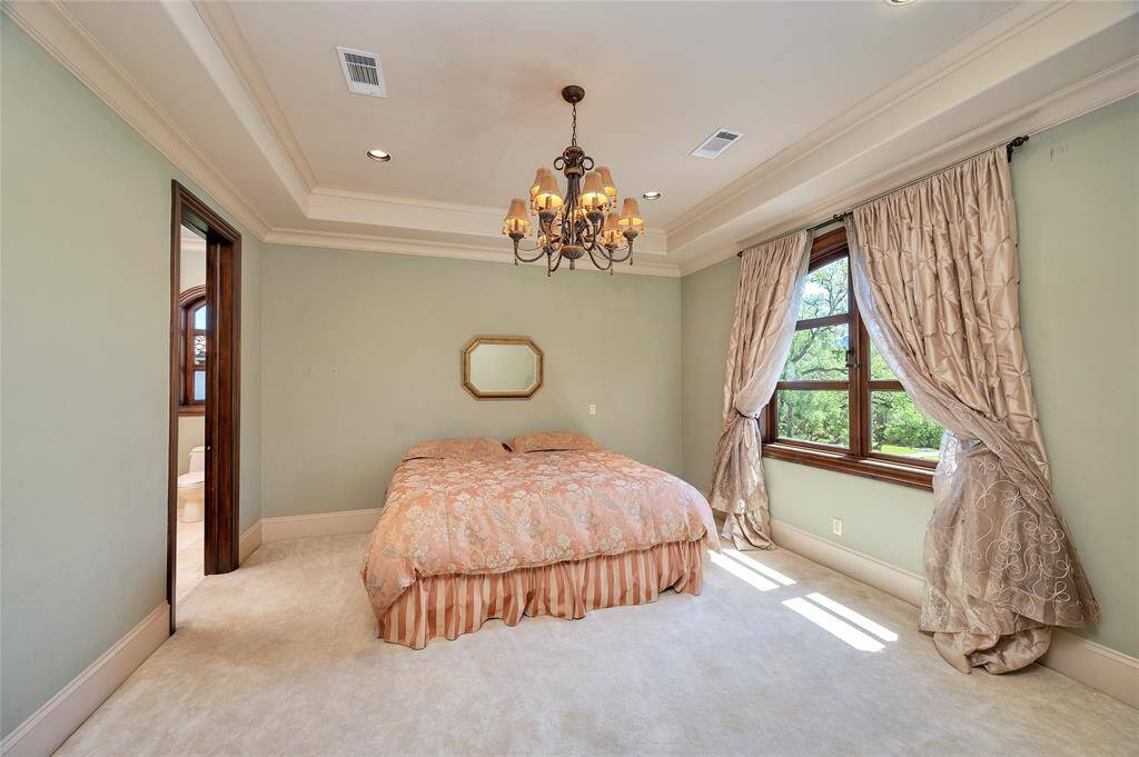 Another secondary bedroom with neutral carpet, baseboards, soft green wall paint, an arched casement window, and tray ceiling with recessed lighting and a bronze statement chandelier. The doorway pictured on the left opens to an ensuite bathroom with a storage closet.