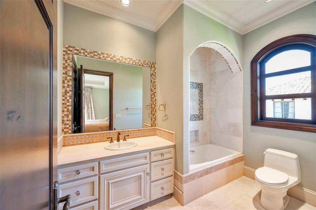 This secondary bathroom off the guest bedroom features travertine tile flooring, a built-in vanity with ample storage, stone countertop, tiled backsplash, glass mosaic tile framed vanity mirror, recessed lighting, an arched window, and Toto toilet. A large soaking tub with a shower and arched opening has a travertine tile surround with a glass mosaic tile inlay and recessed shampoo niche.