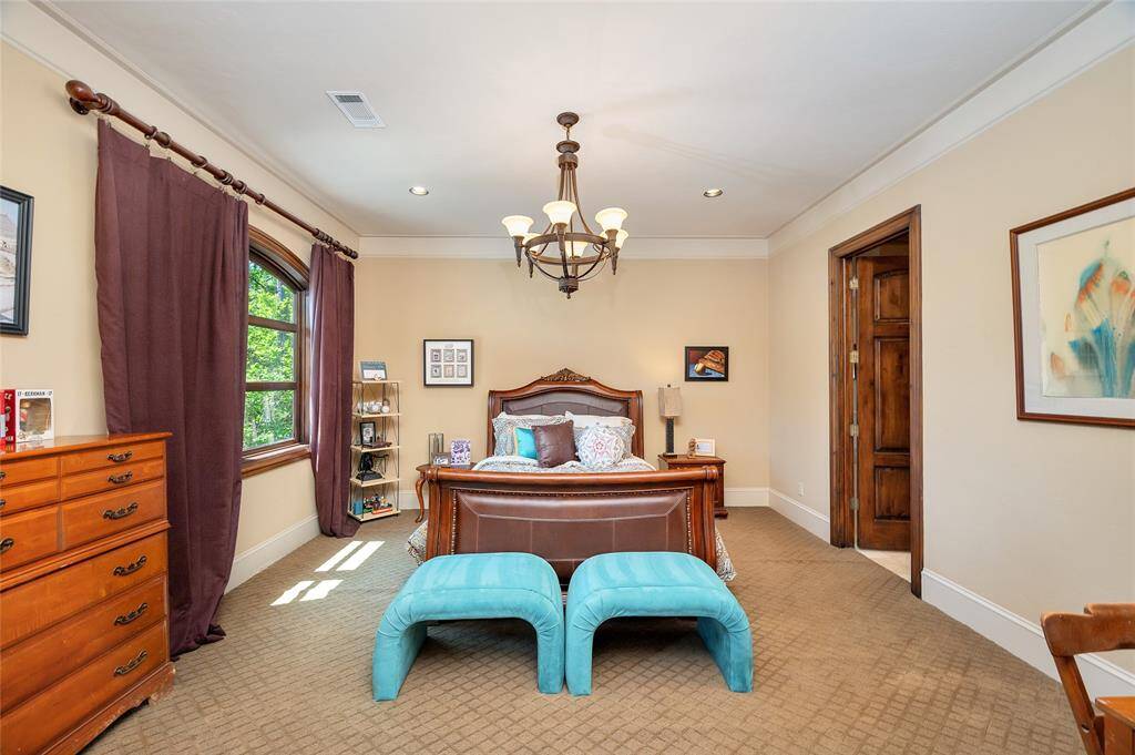 This large secondary bedroom with a sitting area, ensuite bathroom, and walk-in closet features neutral carpet, baseboards, arched windows with elegant draperies, crown molding, recessed lighting, and a bronze chandelier.
