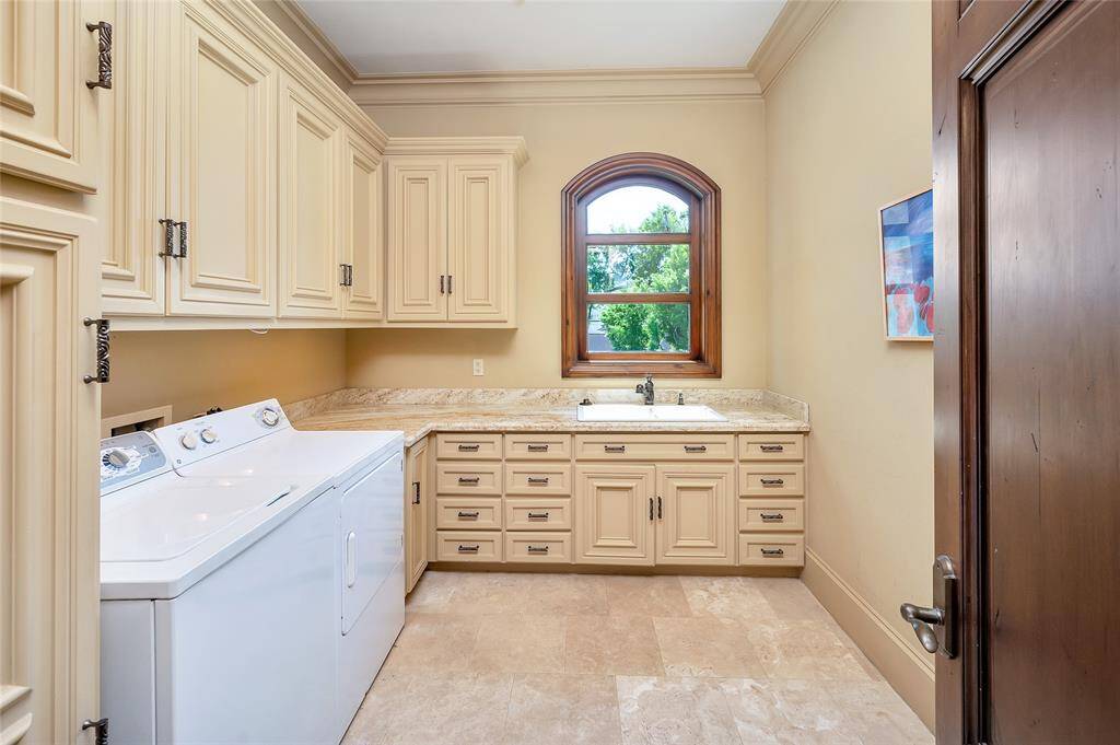 Located on the second floor, the laundry room features custom built-in cabinetry with granite countertops and backsplash, undercabinet lighting, white double basin sink, oil-rubbed bronze faucet with pull-down spray, and bronze cabinet pulls. Additional highlights include an arched window for natural light, wood-painted baseboards and crown molding, travertine tile flooring, and a frosted glass bowl and bronze pendant light fixture.