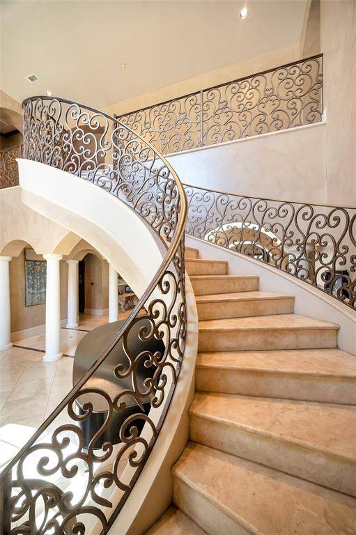 This photo showcases the elegant, curved staircase with  intricate ironwork and travertine slab steps, set in the bright and spacious foyer with high ceilings and dramatic archways.