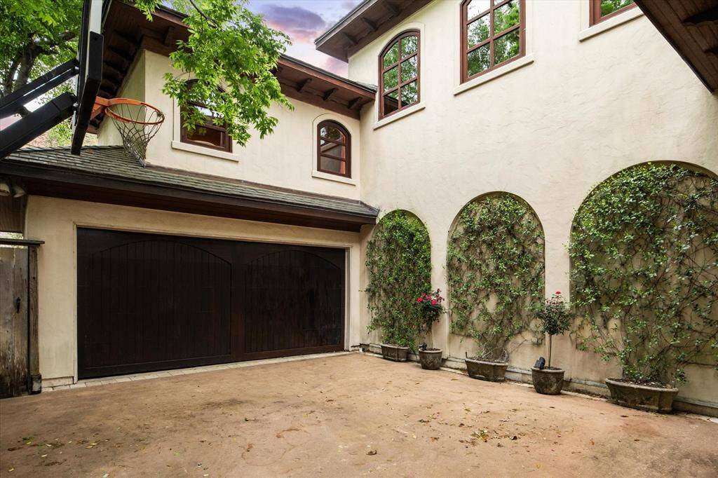 The driveway area doubles as a basketball court and leads to an attached two-car garage with a dark wooden automatic door. Elegant recessed arches covered in greenery add a touch of sophistication.
