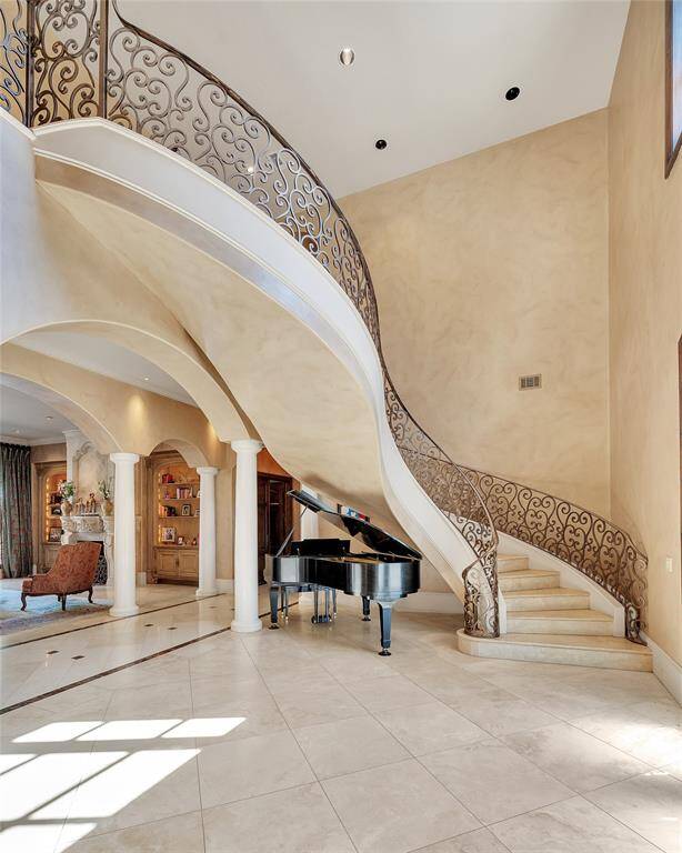 The elegant foyer features a grand, curved floating staircase with ornate wrought iron railings and travertine tile flooring. The space is open and inviting, with 24-ft ceilings and archways leading to additional rooms.