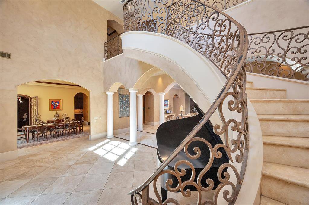 Another view of the spacious foyer highlighting the grand staircase with travertine slab treads and risers and elaborate wrought iron railings. Visible in the background is the formal dining area with an arched entry.