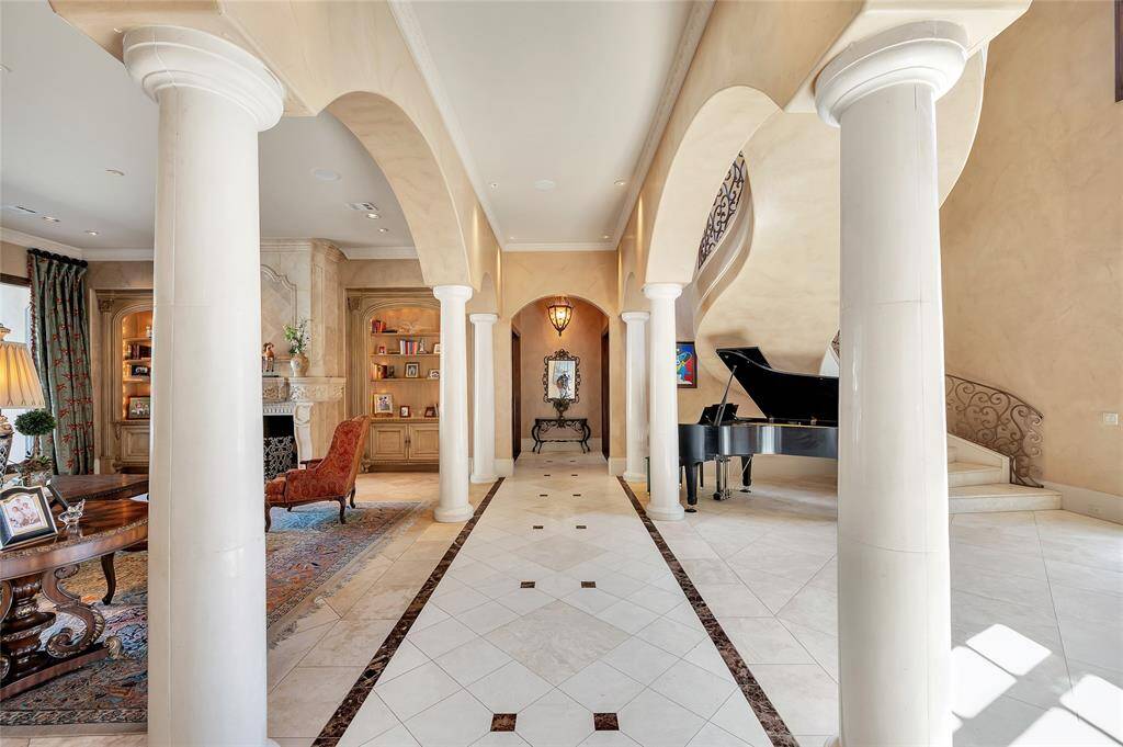 The grand foyer opens to this stunning entry hallway featuring wide archways supported by eight stately limestone columns, allowing an open view of the pool from the front door. Travertine tile flooring with a brown marble tile border and accent inlays enhances the sophisticated ambiance.