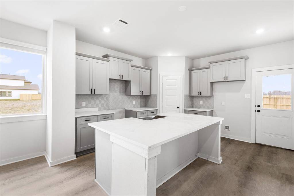 Modern kitchen with gray cabinetry, a large central island, and light countertops. Features a sleek backsplash, recessed lighting, and ample natural light from the window and door.