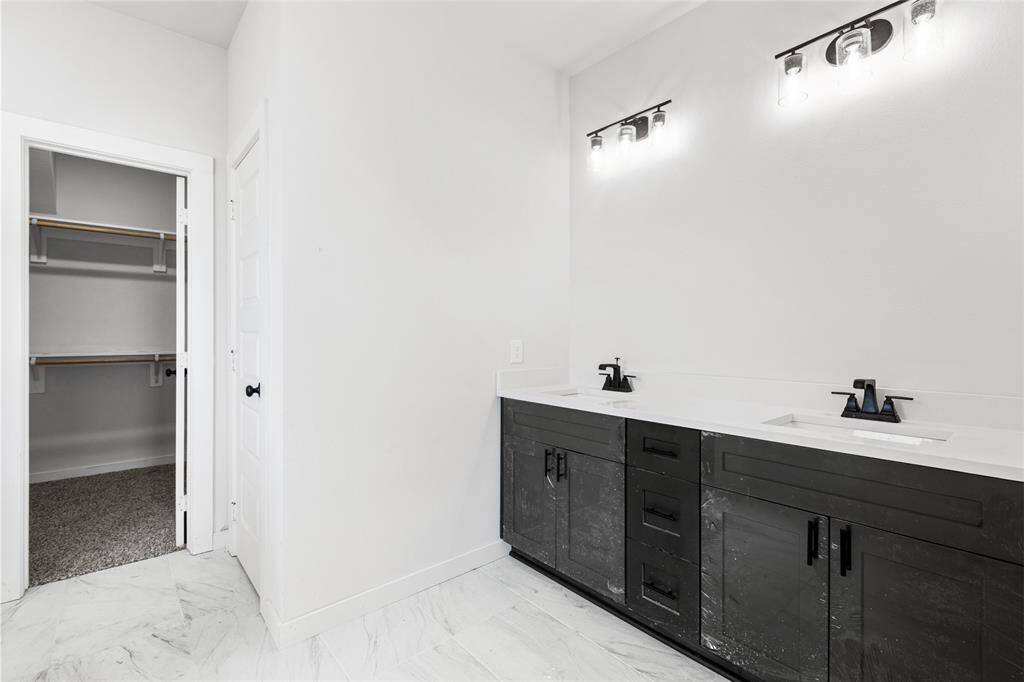 This photo features a modern bathroom with a double vanity, sleek black fixtures, and ample cabinet space. The room has bright lighting and a large walk-in closet with built-in shelving. The flooring is a light-colored tile, adding to the contemporary feel.