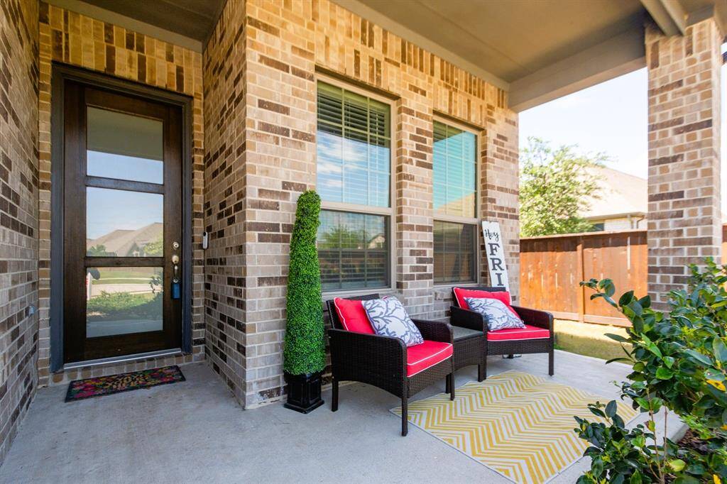This inviting front porch features a cozy seating area set against a warm brick exterior. A modern glass panel door and well-maintained landscaping enhance the welcoming atmosphere.
