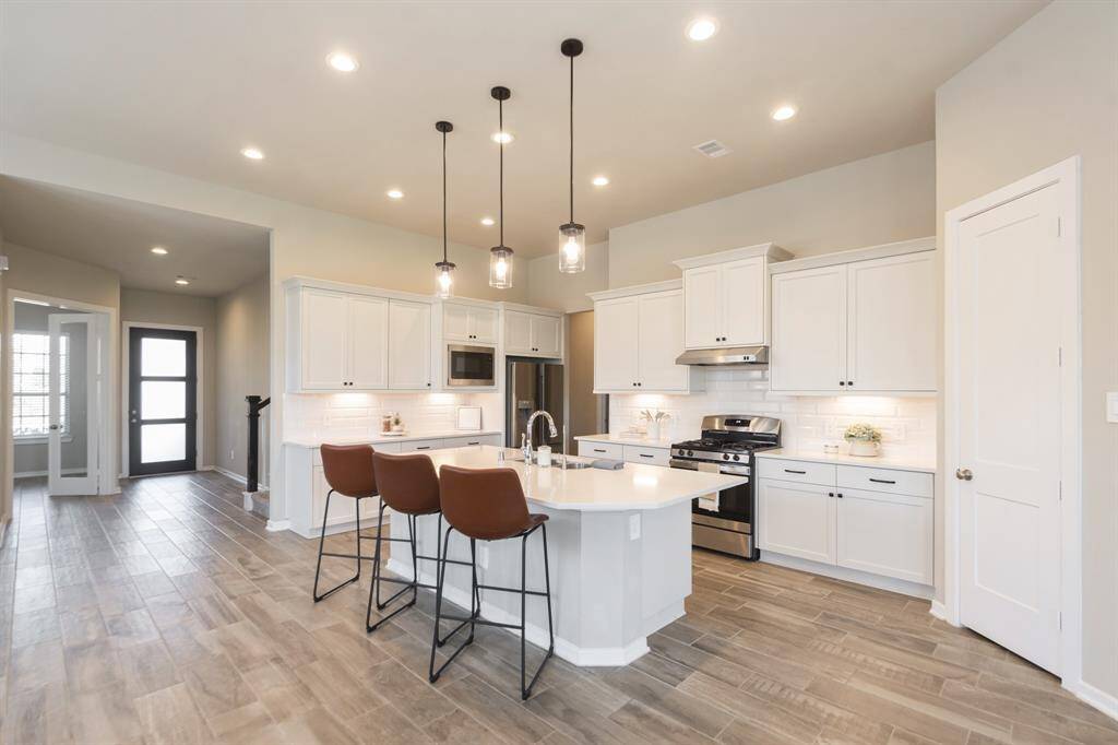 This modern kitchen features a spacious layout with white cabinetry, stainless steel appliances, and a central island with seating. The room is well-lit with recessed lighting and pendant fixtures, complemented by a sleek tile wood look floor. The open design connects to the entryway, enhancing the home's flow.