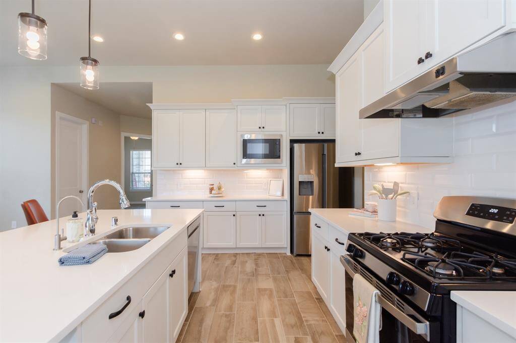 Modern kitchen with sleek white cabinetry, stainless steel appliances, and a large island with a double sink is well-lit with pendant lights and features a gas stove and wood-style flooring, offering a clean and inviting atmosphere.