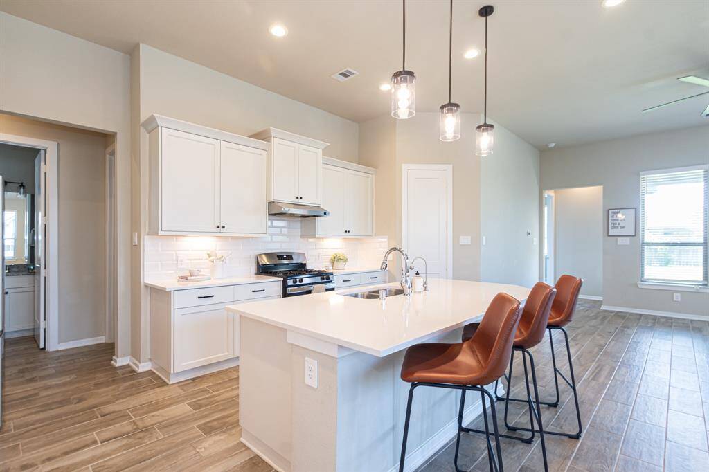 This modern kitchen features white cabinetry, stainless steel appliances, and a spacious island with a sink and seating for three or four. Pendant lighting adds a stylish touch, while wood-look tile flooring extends throughout the open layout, providing a warm and inviting atmosphere.