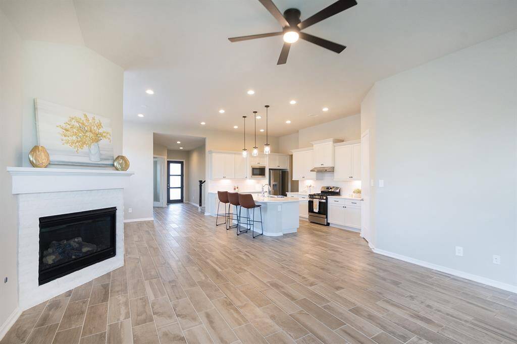 Bright, open-concept living space with modern kitchen featuring a central island and bar seating. The room includes a cozy fireplace, ceiling fan, and wood-look tile flooring, blending functionality with style.