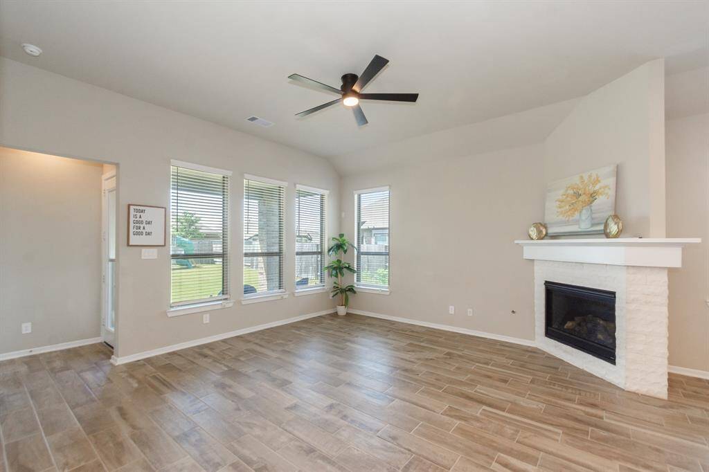 Bright living room with large windows, tiled flooring, and a modern ceiling fan. Features a cozy fireplace with a white mantel, complemented by neutral walls and a view of the backyard.