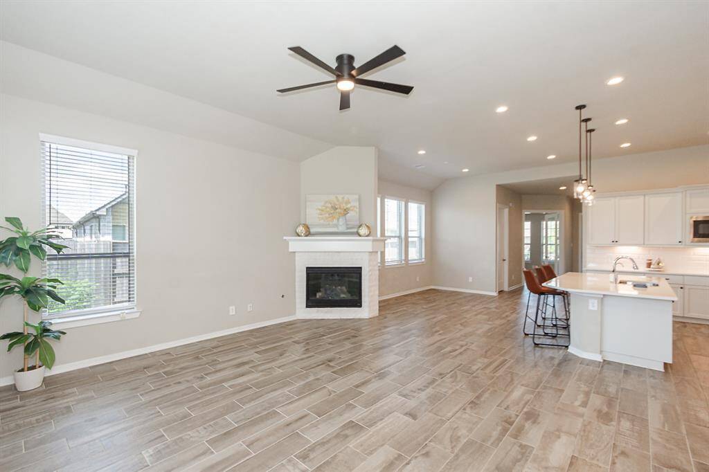 Bright and open living space with wood-look tile flooring, featuring a cozy fireplace and ceiling fan. The room flows seamlessly into a modern kitchen with an island, bar seating, and stylish pendant lighting, perfect for entertaining.