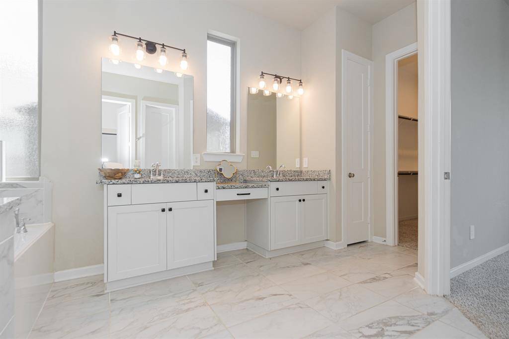 Modern primary bathroom featuring double vanities with granite countertops, framed mirrors, and stylish lighting. It includes a walk-in closet and a glimpse of a luxurious bathtub.