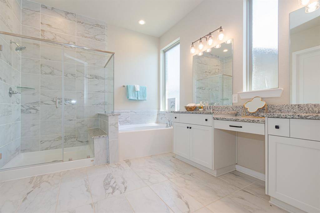 This spacious bathroom features elegant marble tiles, a glass-enclosed shower, and a separate bathtub. The dual-sink vanity offers ample counter space and storage, enhanced by stylish lighting and large windows for natural light.