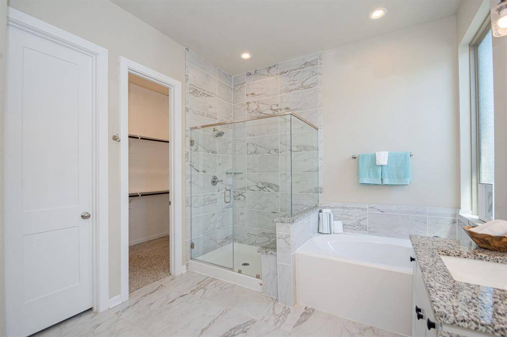 Modern bathroom featuring a glass-enclosed shower, a separate bathtub, granite countertop, and a walk-in closet. Natural light streams through a large window, enhancing the bright, elegant design.