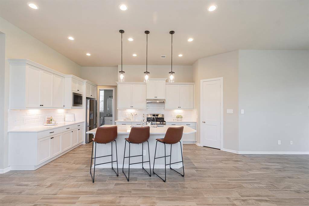 This modern kitchen features sleek white cabinetry, stainless steel appliances, and a stylish island with pendant lighting. The open layout and wood-look tile flooring create a spacious and inviting atmosphere.