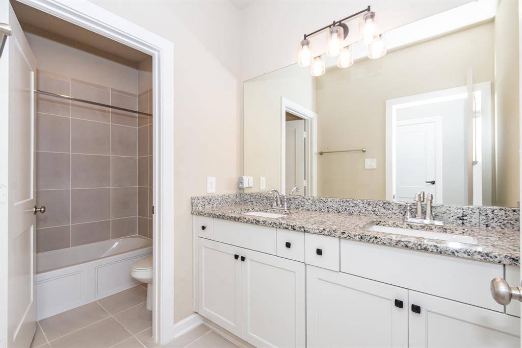 This upstairs full bathroom features a modern design with a double vanity topped with granite, a large mirror, and stylish lighting. It includes a shower-tub combo with tiled walls, and neutral tones create a clean, spacious feel.