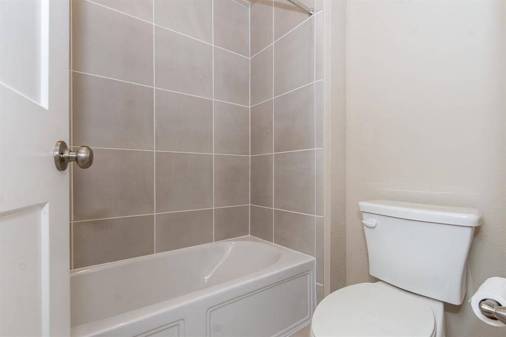 This bathroom features a modern design with neutral-toned tiles surrounding a bathtub, complemented by a white toilet and light walls for a clean, fresh look.