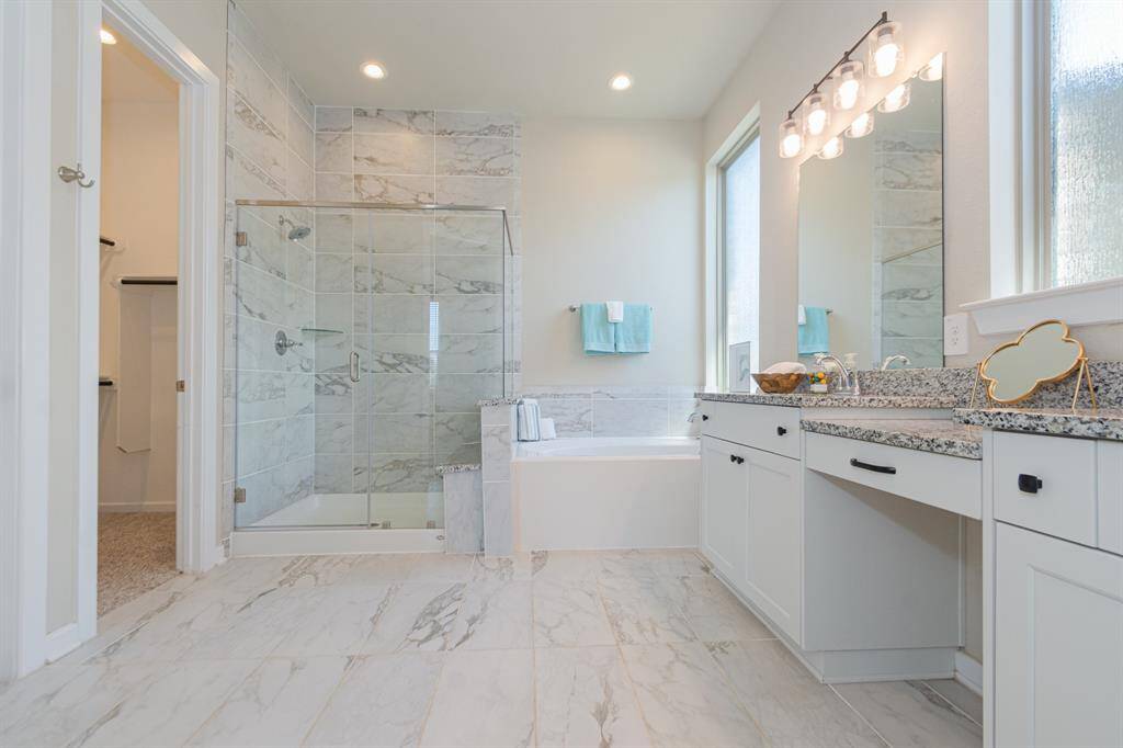 This primary bathroom features a modern design with marble tile flooring, a spacious glass-enclosed shower, a large soaking tub, and dual vanities with granite countertops. Natural light floods the space, highlighting its elegant and clean aesthetic.
