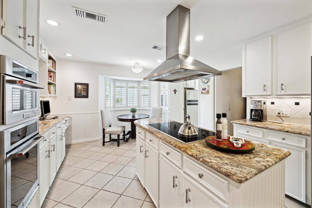 This kitchen features a spacious layout with white cabinetry, granite countertops, and a central island with a cooktop and modern range hood. It includes built-in ovens, a cozy breakfast nook by a bay window, and plenty of natural light.